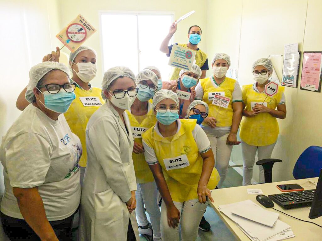 Diversos colaboradores estão bem próximos posando de frente para a câmera. Todos usam uniforme, touca e máscara. Alguns seguram nas mãos placas com frases e desenhos relacionados à proibição do uso de adorno na unidade.