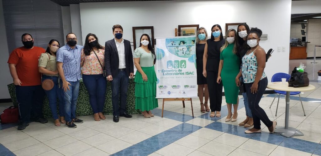 Foto com 11 colaboradores do ISAC que participaram do encontro. Eles estão em uma sala grande. Atrás deles há uma mesa com toalha verde estampada e uma parede branca onde estão três quadros com moldura marrom. As pessoas estão lado a lado de frente para a câmera. Todas usam máscara. Entre elas há um banner do evento. Quatro estão de lado do banner e as outras seis do outro lado. Há uma mochila vermelha no chão, ao lado da primeira pessoa do lado esquerdo.