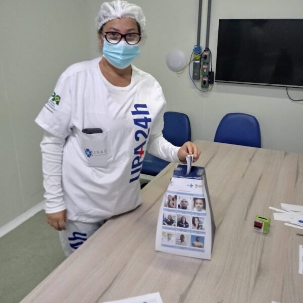 Uma mulher está de frente para a câmera. Ela usa máscara, uniforme branco da UPA, touca e óculos. Ela aparece colocando a cédula de votação na urna. Na mesma mesa onde está a urna, há vários papeis espalhados, além de caneta e um carimbo.