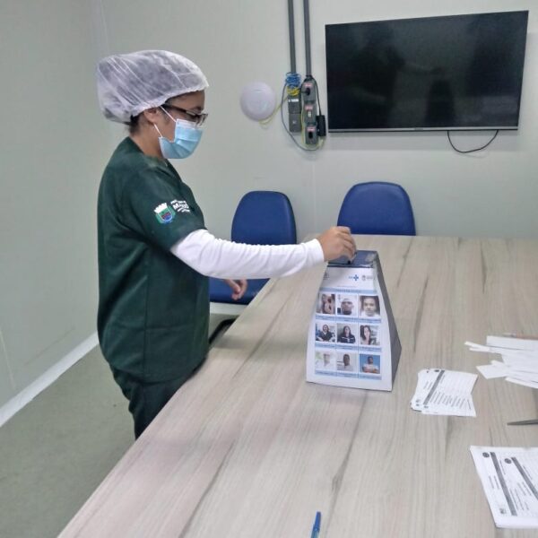 Uma mulher está de perfil para a câmera. Ela usa máscara, uniforme verde, touca e óculos. Ela aparece colocando a cédula de votação na urna. No fundo há uma televisão suspensa na parede.