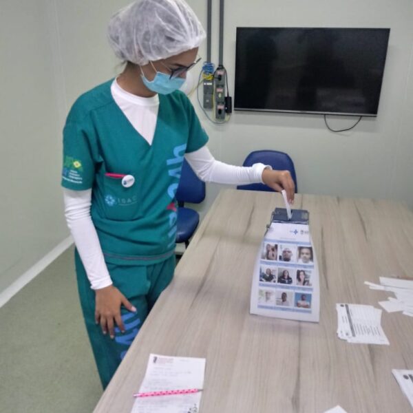 Uma mulher está de lado. Ela usa máscara, uniforme verde, touca e óculos. Ela aparece colocando a cédula de votação na urna. Na frente dela, há uma mesa onde estão a urna e vários papeis espalhados. No fundo da sala há uma televisão suspensa na parede.