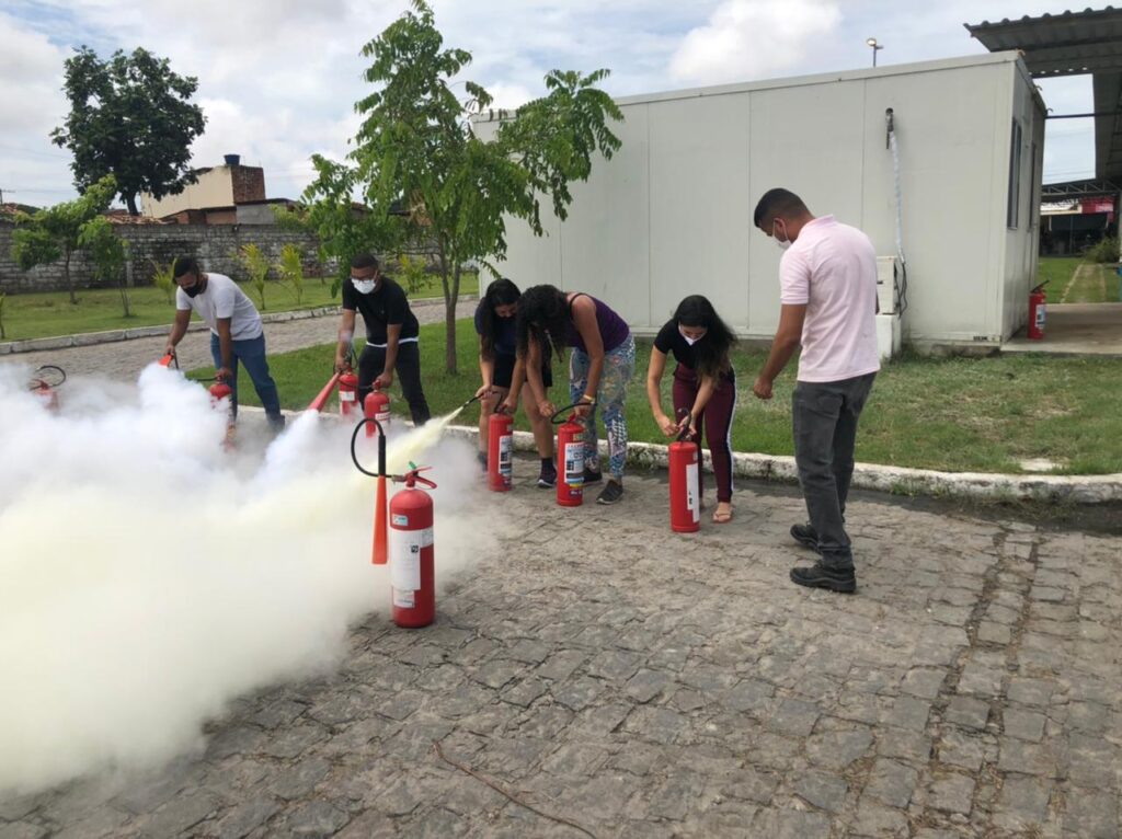 A imagem mostra seis colaboradores da UPA Trapiche da Barra na área externa da unidade fazendo a simulação do uso de extintores de incêndio.