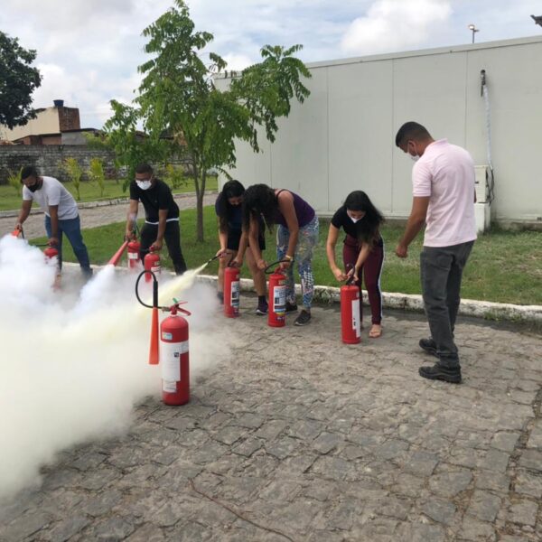 A imagem mostra seis colaboradores da UPA Trapiche da Barra na área externa da unidade fazendo a simulação do uso de extintores de incêndio.