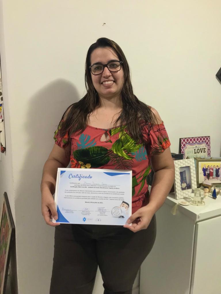 Mulher está de pé, posando para câmera e segundando o certificado da ONA 2. Ela usa óculos de grau e sorri para câmera.