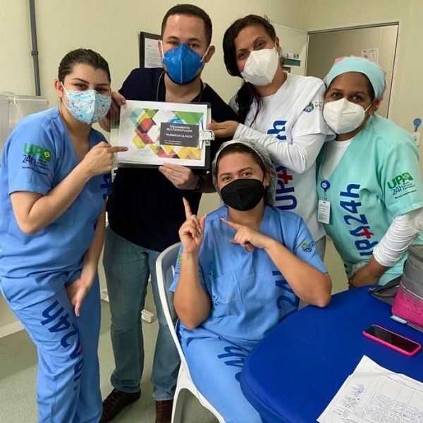 Quatro pessoas estão de pé e uma sentada posando para câmera. Todas usam máscara e uma delas segura um cartaz.